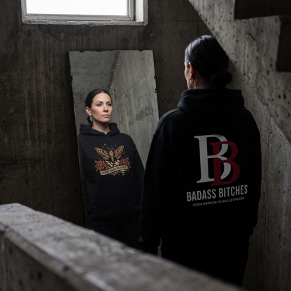 Women wearing a Ashes Made Me Holy hoodie with 'Badass Bitches' logo in a dimly lit room.