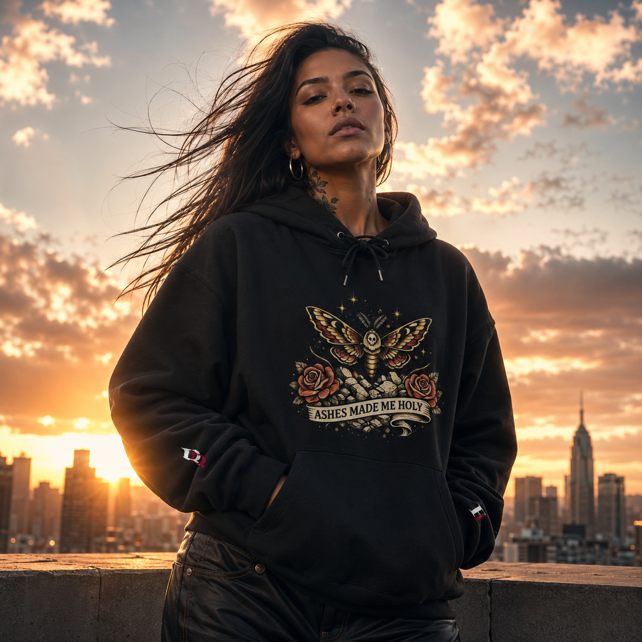Women wearing a Ashes Made Me Holy black hoodie with a butterfly design against a city skyline at sunset.