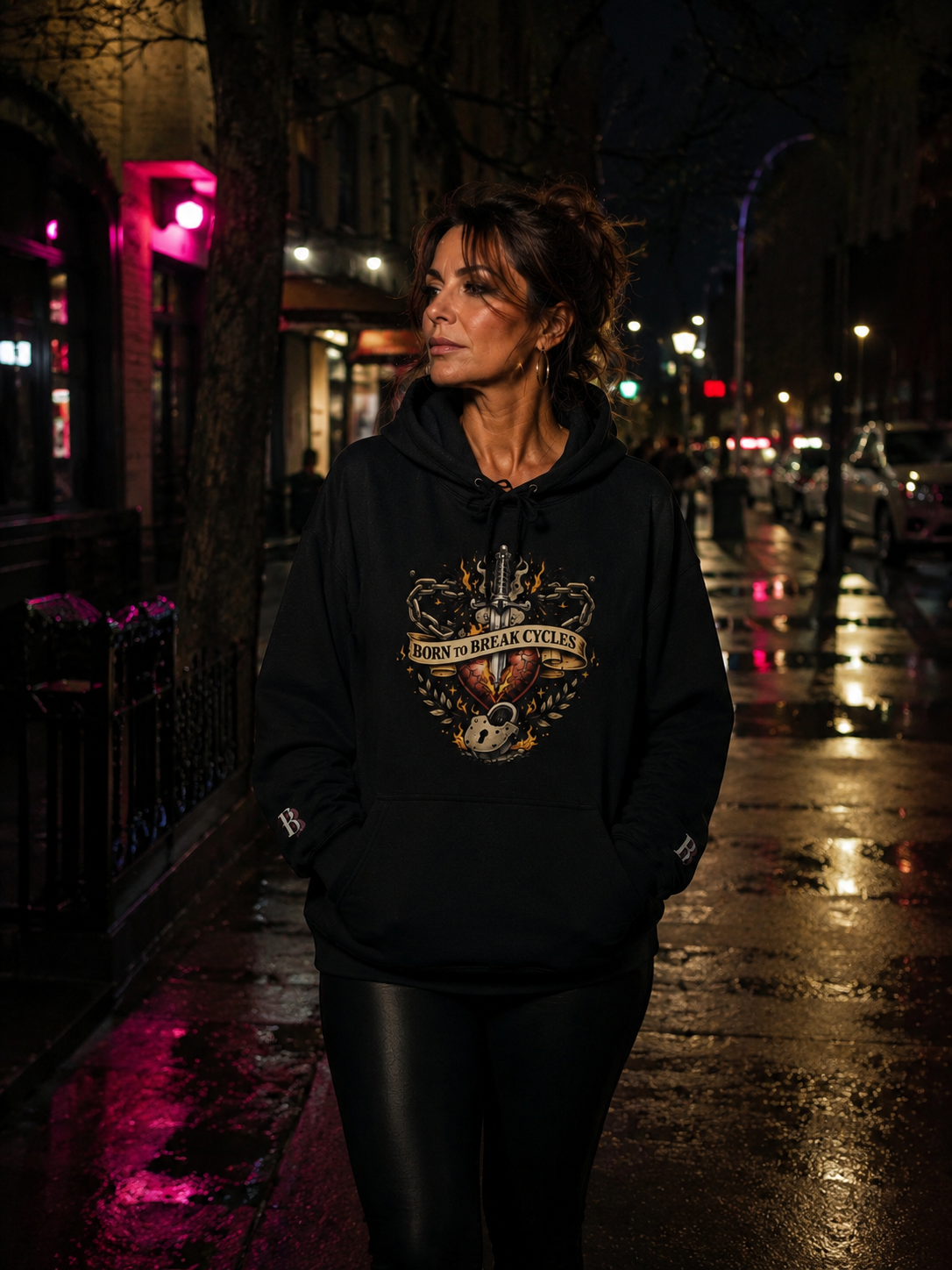 Women wearing a Born To Break Cycles black hoodie with a graphic design on a city street at night.