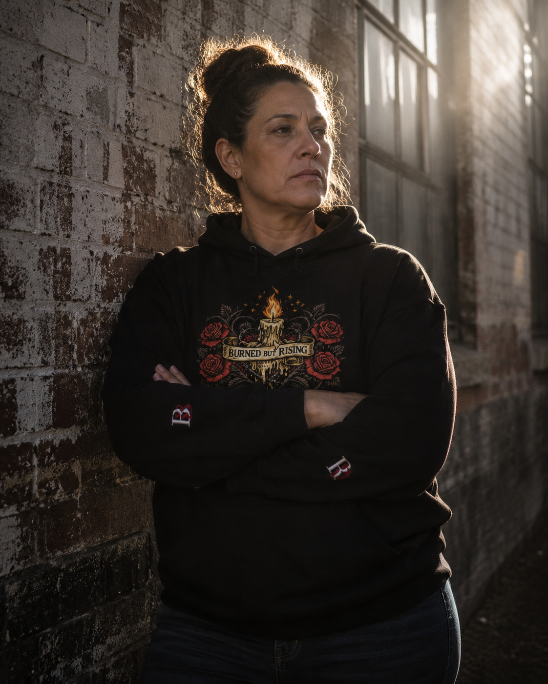 Women wearing a Burned But Rising black hoodie with a graphic design, standing against a textured brick wall.