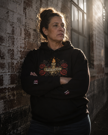 Women wearing a Burned But Rising black hoodie with a graphic design, standing against a textured brick wall.