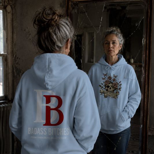Woman wearing aI Walked Through Fire I Didn't Burn blue hoodie with a Badass Bitches logo, standing in a dilapidated room.
