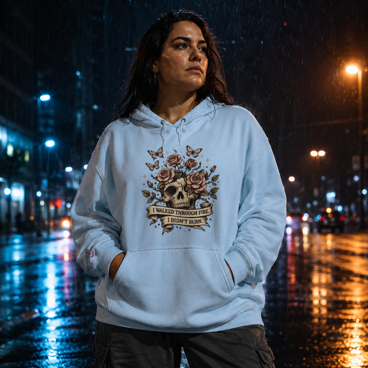 Women wearing a I Walked Through Fire I Didn't Burn blue hoodie with a skull and roses design on a city street at night.