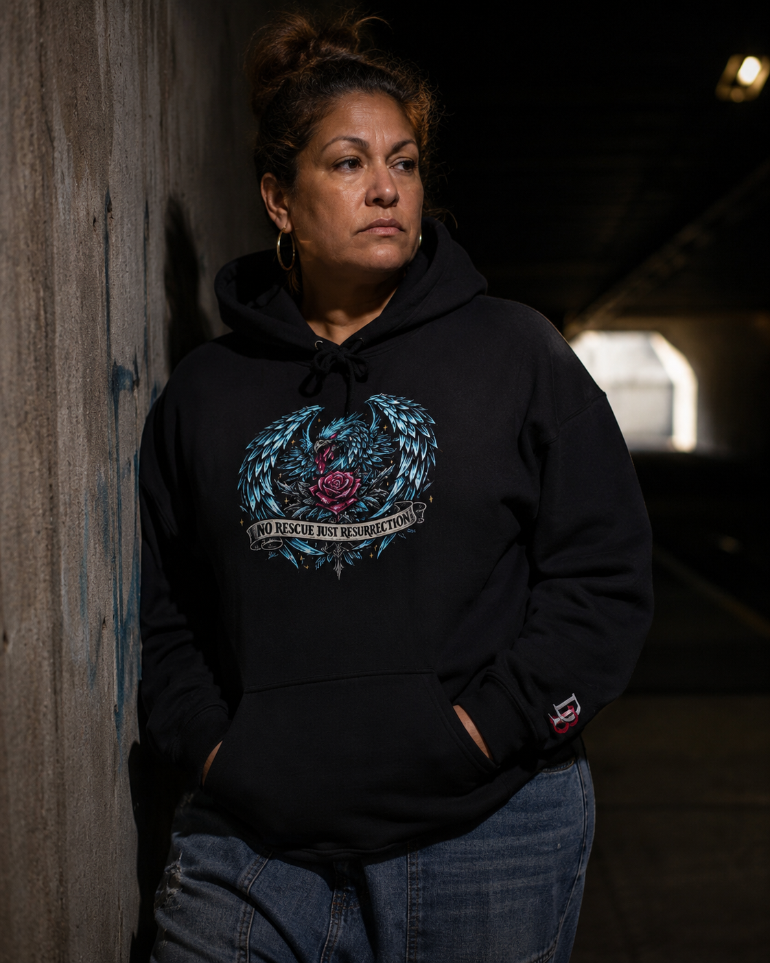 Women wearing a No Rescue Just Resurrection black hoodie with a graphic design, standing against a dark wall.