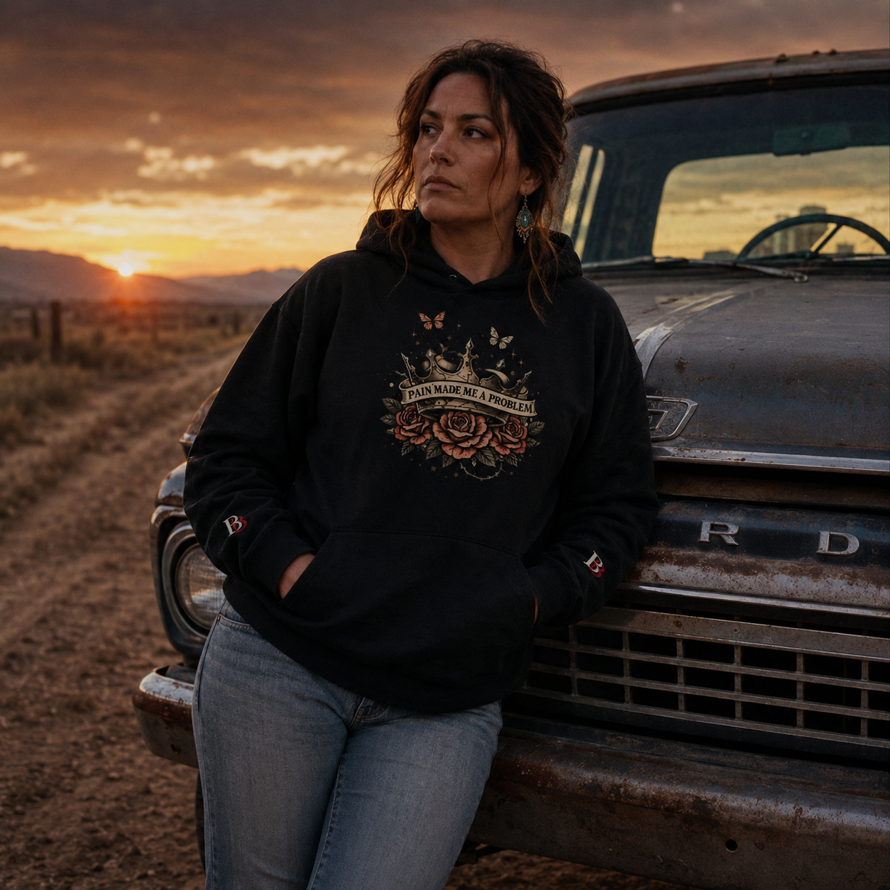 Woman in a "Pain Made Me A Problem" black hoodie leaning against an old truck with a sunset in the background