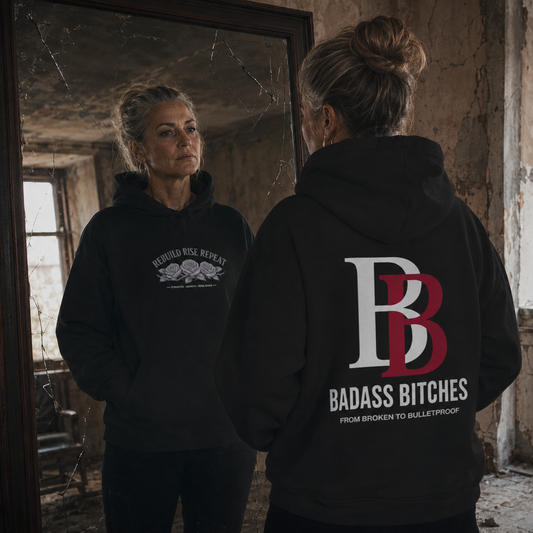 Women wearing a "Rebuild Rise Repeat" black hoodie with a logo and text, standing in a dilapidated room.