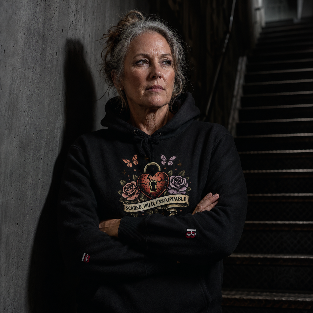 Woman wearing a Scared Wild Unstoppable black hoodie with a design, standing against a concrete wall.