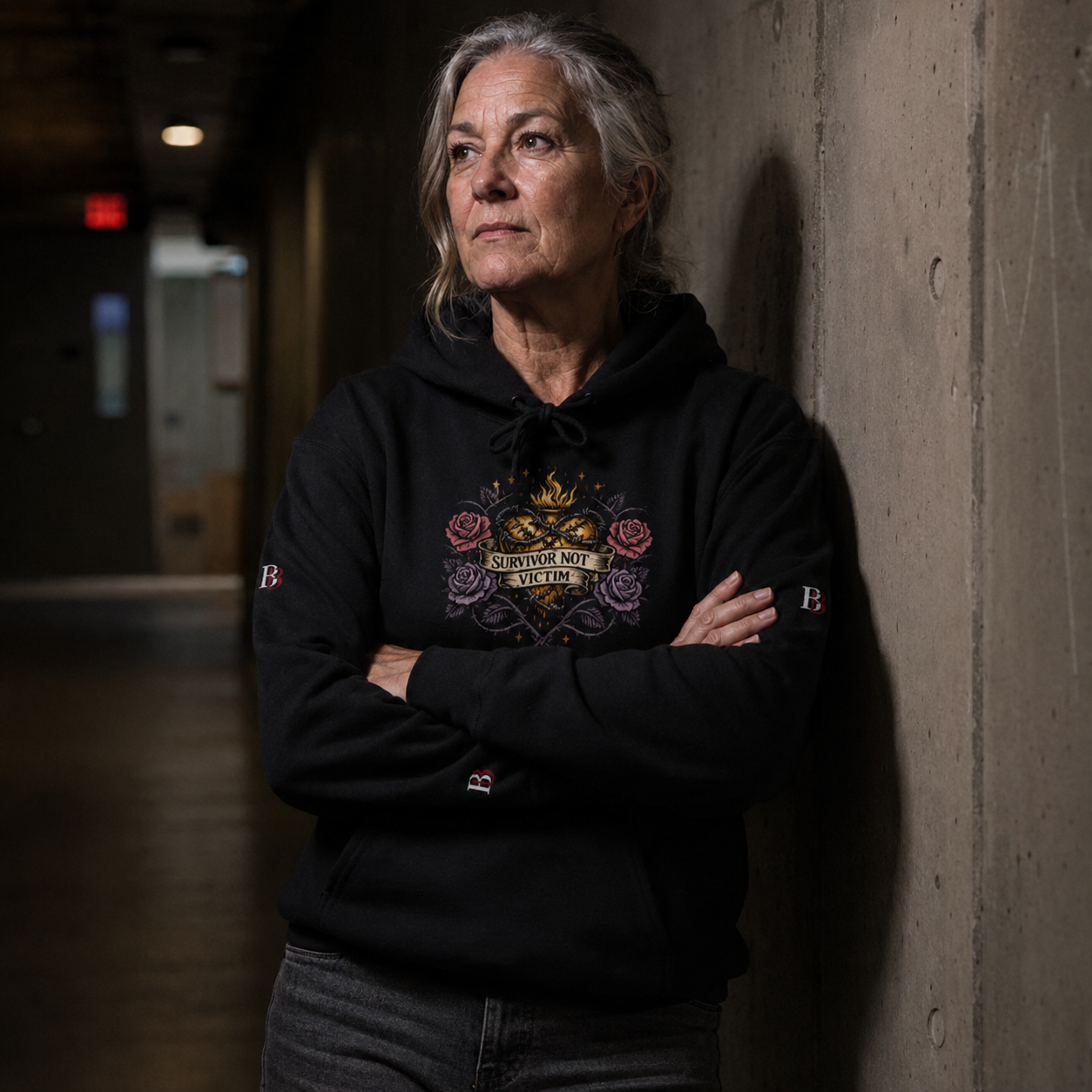 Women wearing a "Survivor Not Victim" black hoodie with a visible "Badass Bitches" logo, standing against a concrete wall.