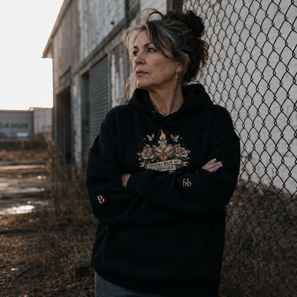 Woman wearing a "Trauma Made Me Stronger" black hoodie with a graphic design, standing in front of a chain-link fence.