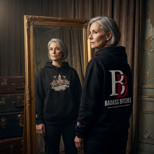 Woman wearing a hoodie with "Trauma Made Me Stronger" graphic and 'Badass Bitches' logo standing in front of a mirror.