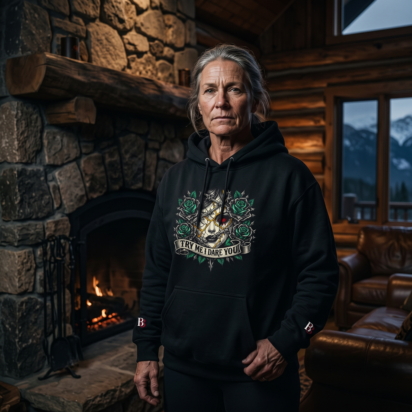 Women wearing a Try Me I Dare You black hoodie with a graphic design in a cozy room with a stone fireplace and mountain view.