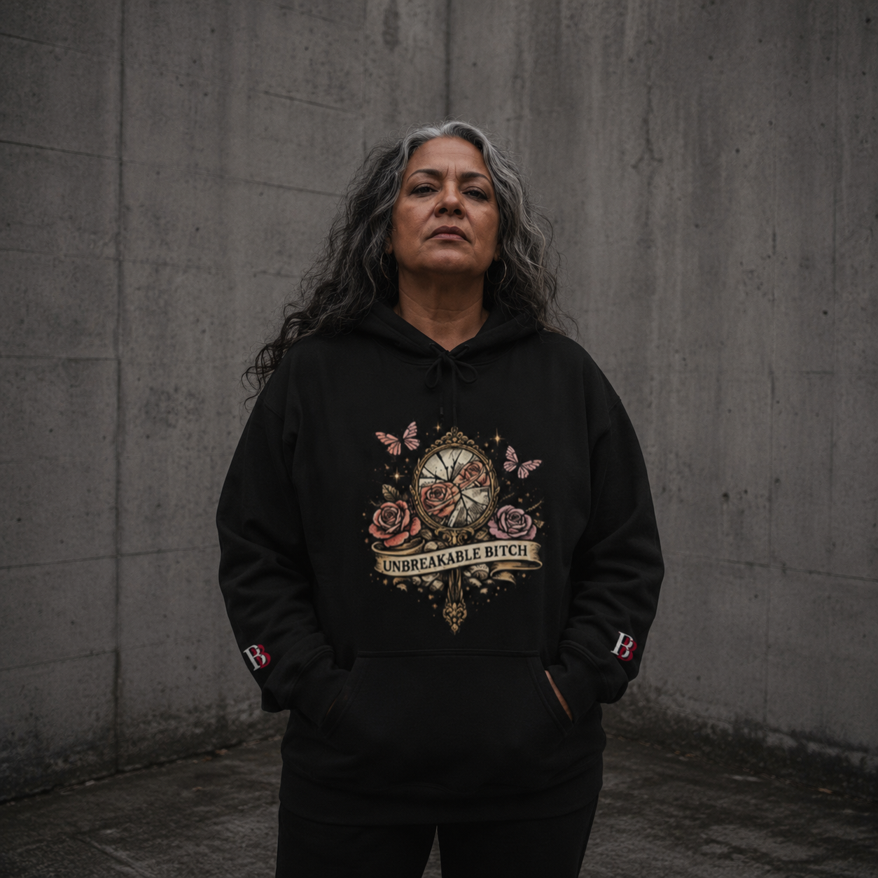 Women wearing a Unbreakable Bitch black hoodie with a graphic design against a gray wall.