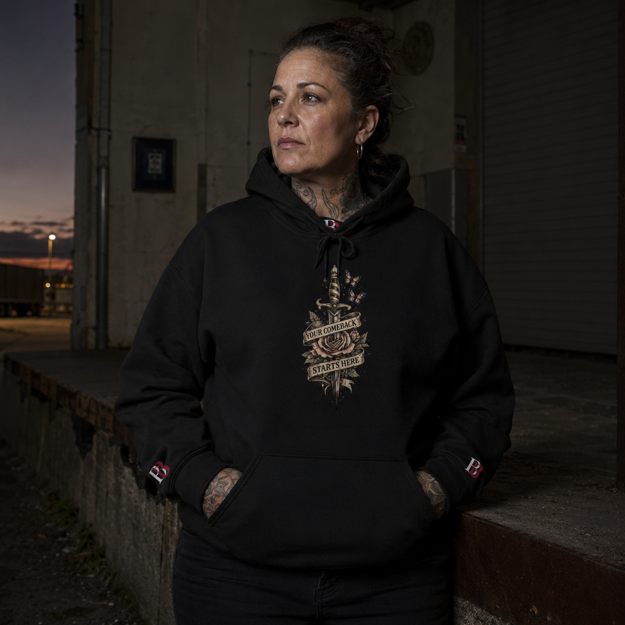 Person wearing a "Your Comeback Starts Here" black hoodie with a "Badass Bitches" logo, standing against a dark background.